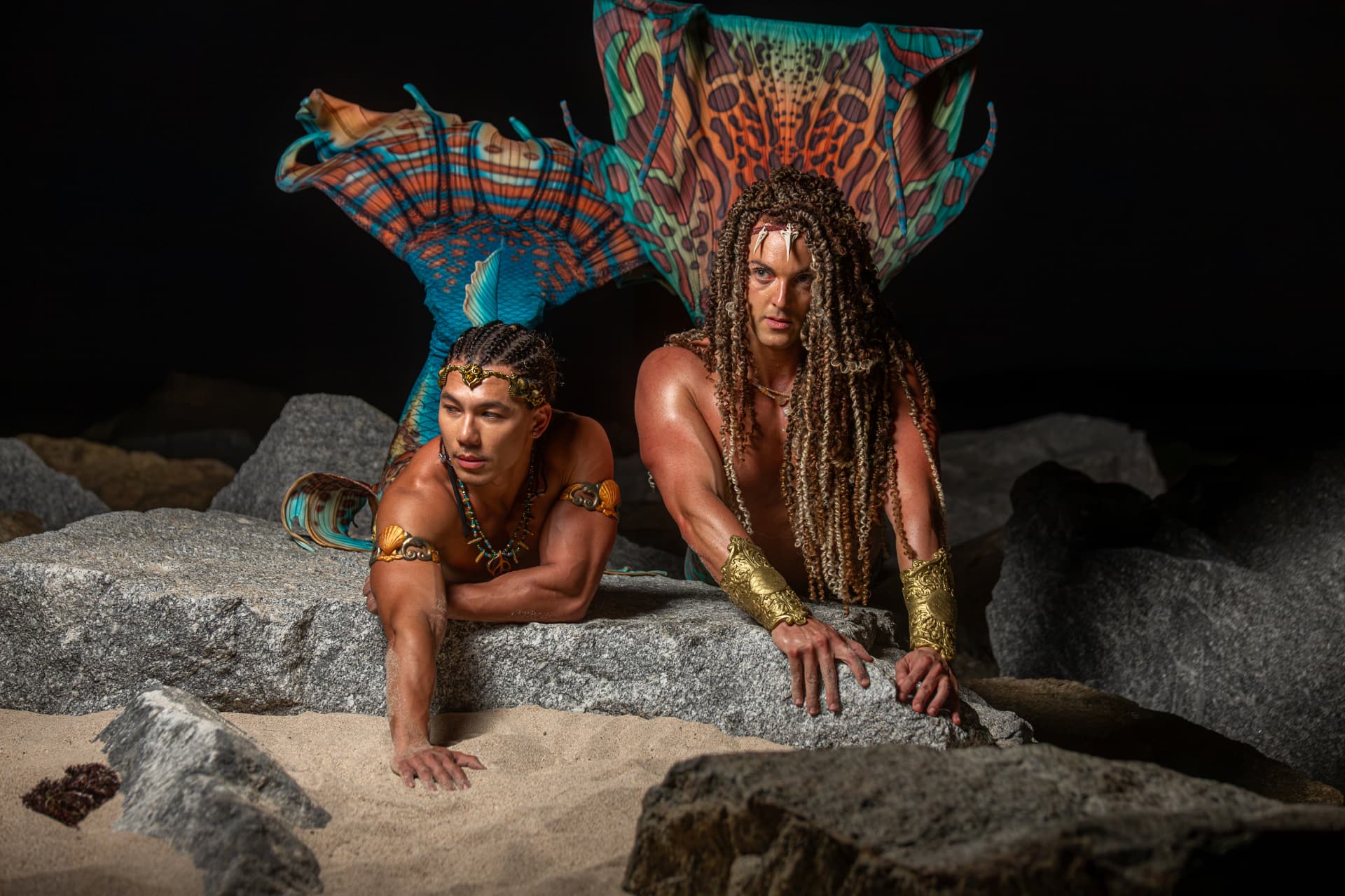 Prince Pōhaku and a fellow merman resting on coastal rocks at night, tails raised
