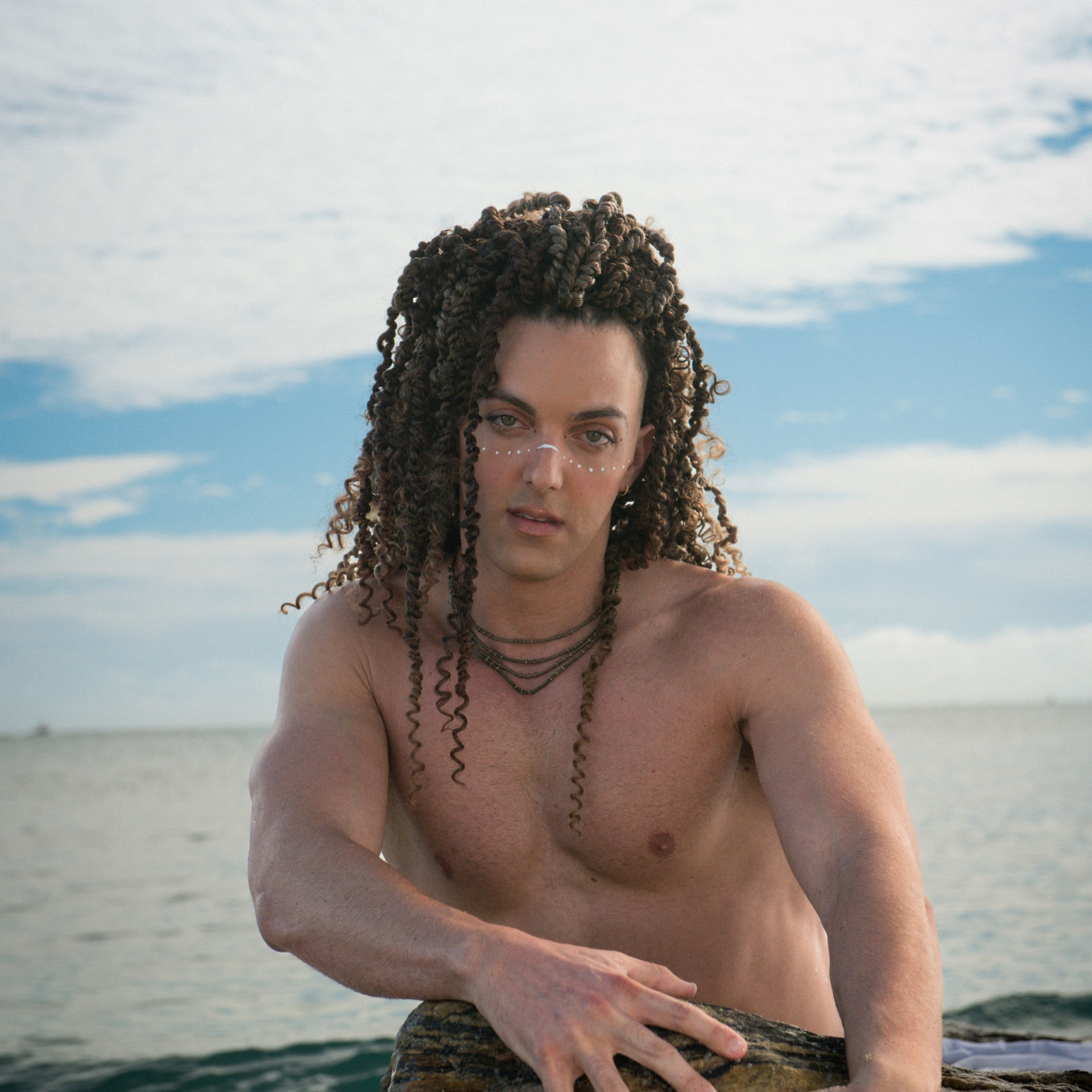 Prince Pōhaku leaning on a rock at the water's edge, white paint across his nose and cheeks