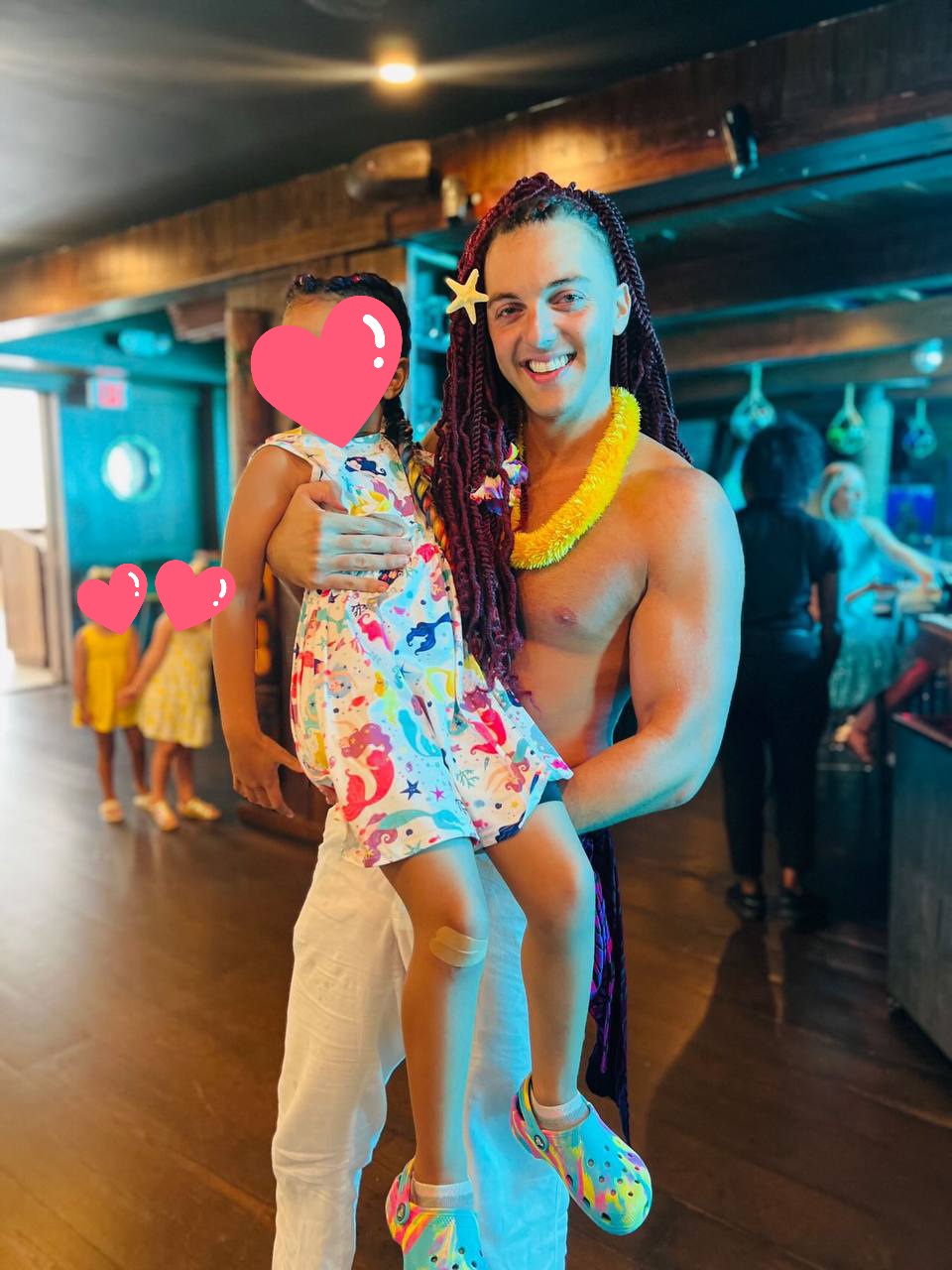 Prince Pōhaku holding a smiling child at a celebration, yellow flowers around his neck, a magenta and violet fin at his hip